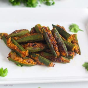 How To Make Bharwa Bhindi Bharwa Bhindi on a white bowl with some coriander leaves on the background kept on a wooden surface with a spoon