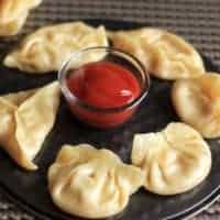 How To Make Cheese Corn Veg Momos Recipe Cheese corn momo on a black round plate with some tomato sauce in the center putted on a dark coloured mat |
