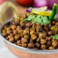 How to make Dry Kala Chana Recipe Dry kala chana on a deep vessel with coriander leaves and green chilli on top with a slice of lemon and some onion rings and light background with onion and tomato in a basket.