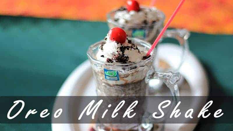 Oreo-Milk-Shake on a white surface with 2 glasses of milkshake decorated with some heavy cream with some oreo biscuit crush and a cherry on the top with a strow in it |