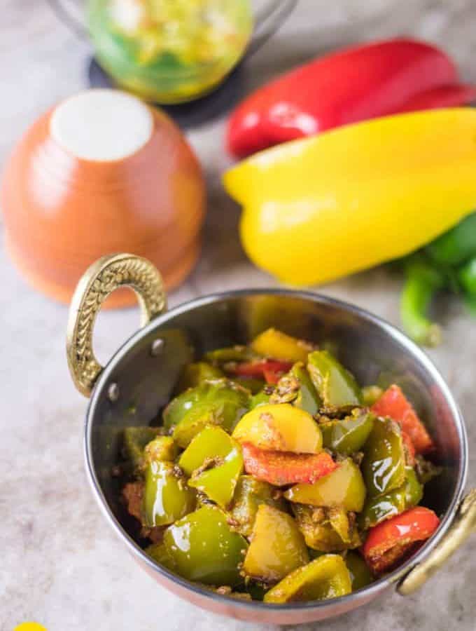 Achari shimla mirch on a deep vessel with some capsicum and bell pepper in the background with a clay pot kept on a white marble |