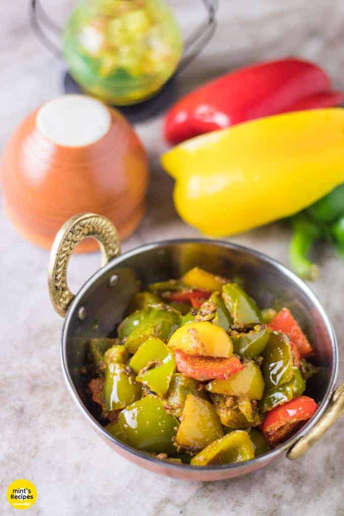 Achari shimla mirch on a deep vessel with some capsicum and bell pepper in the background with a clay pot kept on a white marble |