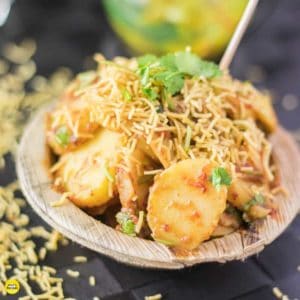 Aloo chaat on a disposable plate garnished with some sev and coriander leaves with a toothpick on a dark mat sprinkles with some sev |
