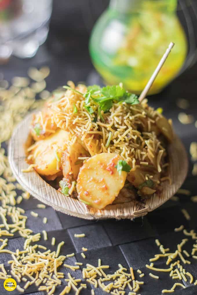 Aloo chaat on a disposable plate garnished with some sev and coriander leaves with a toothpick on a dark mat sprinkles with some sev |