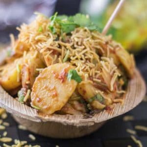 Aloo chaat on a disposable plate garnished with some sev and coriander leaves with a toothpick on a dark mat sprinkles with some sev | www.mintsrecipes.com |