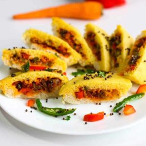 Bread dhokla sandwich on a white plate cutted into pieces in half and tempering of mustard seed and chilli with few strings of curry leaves
