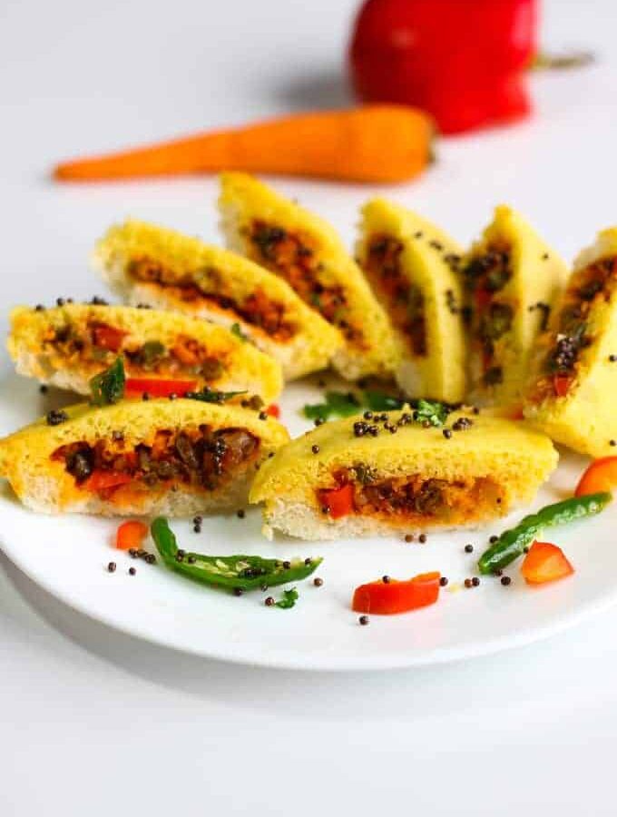 Bread dhokla sandwich on a white plate cutted into pieces in half and tempering of mustard seed and chilli with few strings of curry leaves