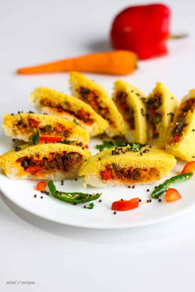 Bread dhokla sandwich on a white plate cutted into pieces in half and tempering of mustard seed and chilli with few strings of curry leaves 
