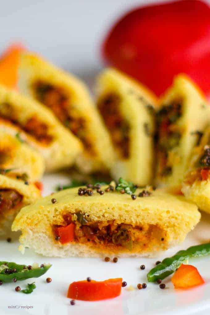 Bread dhokla sandwich on a white plate cutted into pieces in half and tempering of mustard seed and chilli with few strings of curry leaves 