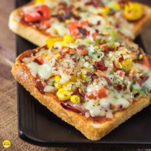 Bread pizza on a black plate with some pieces of bread pizza garnished with some oregano and chilli flakes kept on wooden surface