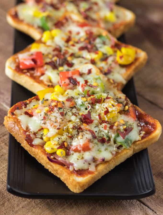 Bread pizza on a black plate with some pieces of bread pizza garnished with some oregano and chilli flakes kept on wooden surface