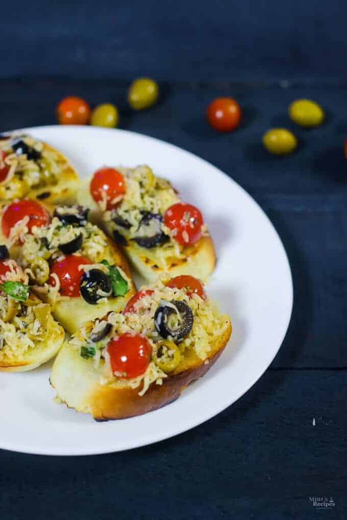 Bruschetta on a white plate of bruschetta garnished with grated cheese kept on a dark wooden surface with some red and green olives | 