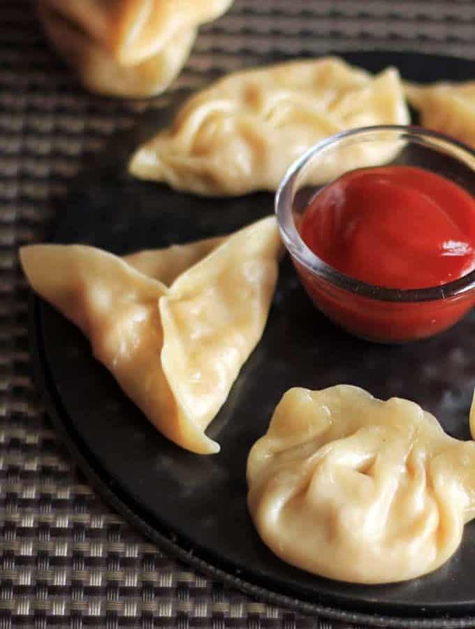Cheese corn momo on a black round plate with some tomato sauce in the center putted on a dark coloured mat |