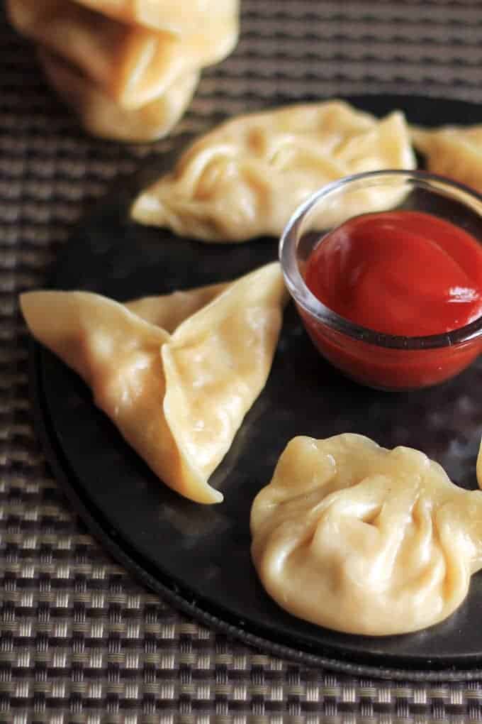 Cheese corn momo on a black round plate with some tomato sauce in the center putted on a dark coloured mat |