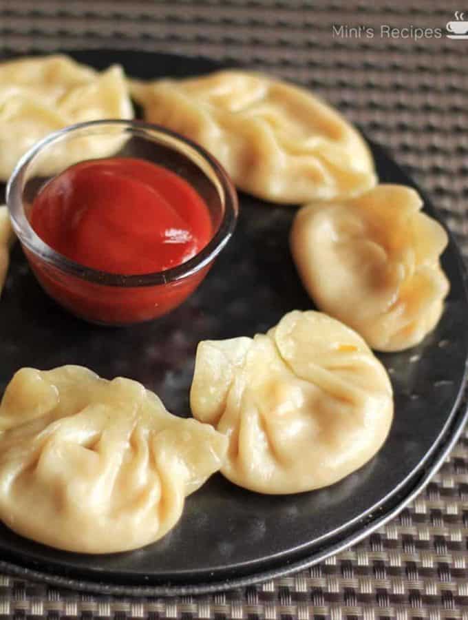 Cheese corn momo on a black round plate with some tomato sauce in the center putted on a dark coloured mat |