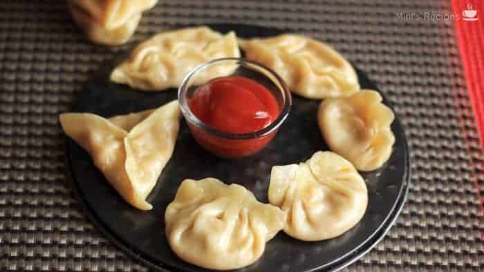 Veg momos on a white plate with some red chilli sauce in the center with a grey surface in background |