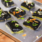 Chocolate Barfi on a steel tray with some pieces of chocolate khoya barfi with some garnishing of pista, and almonds kept on a wooden surface |