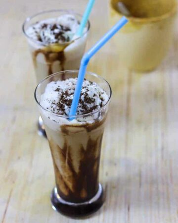 Cold Coffee with Icecream on a glass with some garnishing of chocolate syrup and a strow in it and on the background another glass of coffee ice-cream kept on a wooden surface |