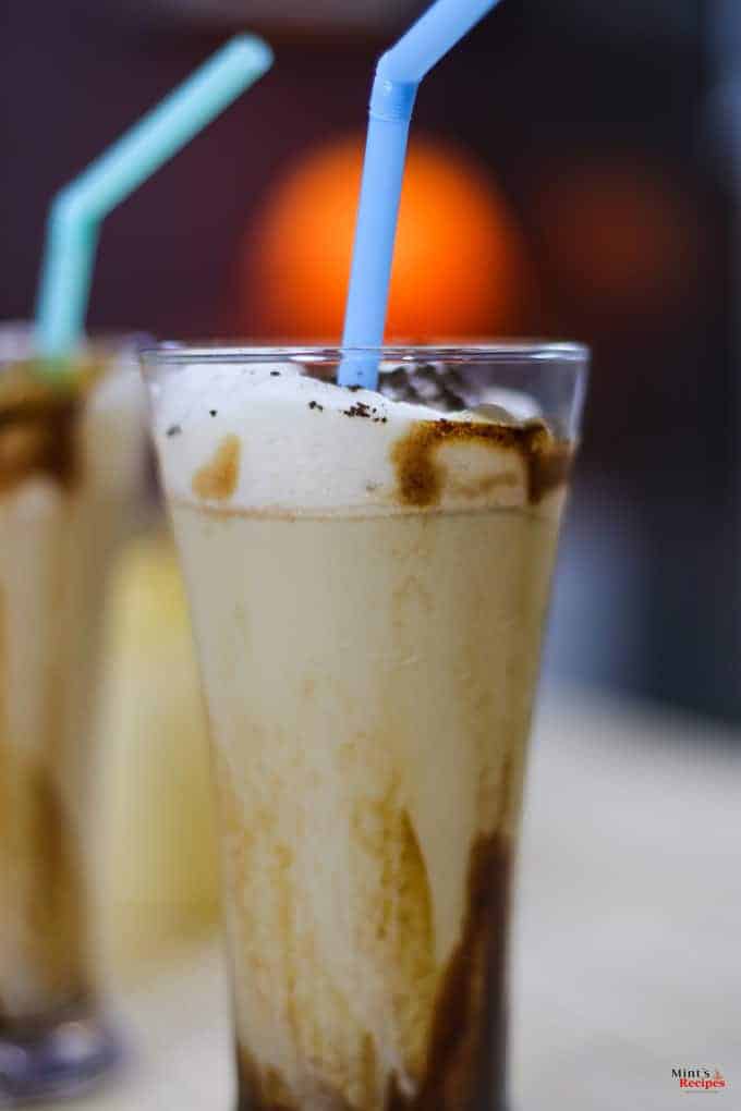 Cold Coffee with Icecream on a glass with some garnishing of chocolate syrup and a strow in it and on the background another glass of coffee ice-cream kept on a wooden surface |