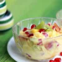 Fruit custard on a transparent bowl putted on a whie plate with some pomogranate on a green matress|