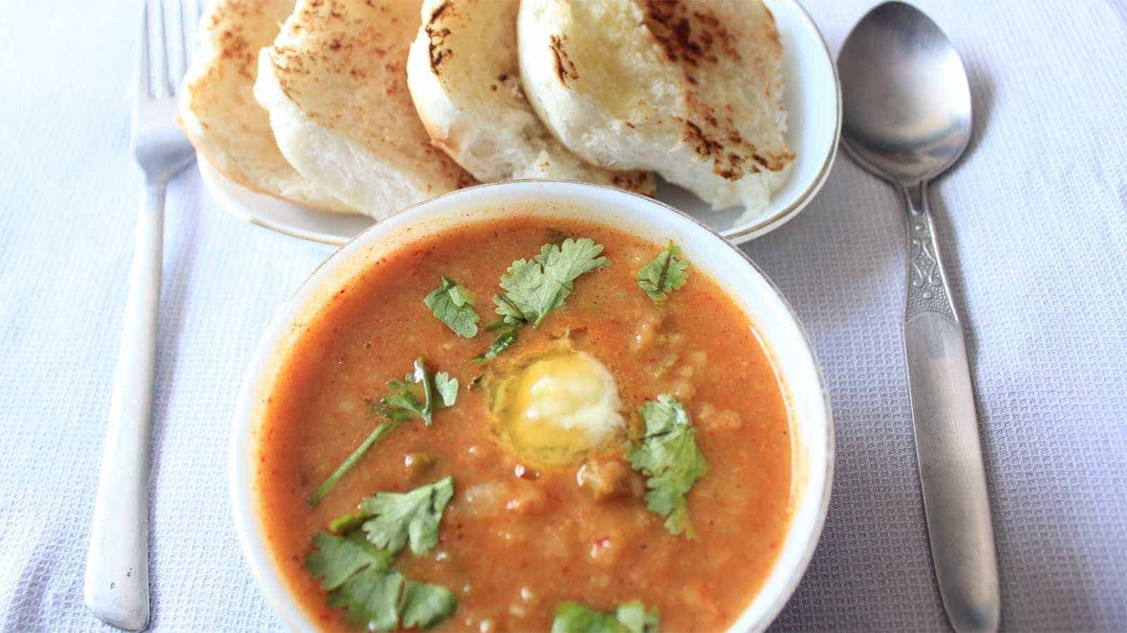 Indian Pav Bhaaji on a white surface wit for pav pieces on a white tray and a bowl full of bhaji with little butter and garnished with some coriander leaves and a spoon,fork in both side oh pav bhaji |