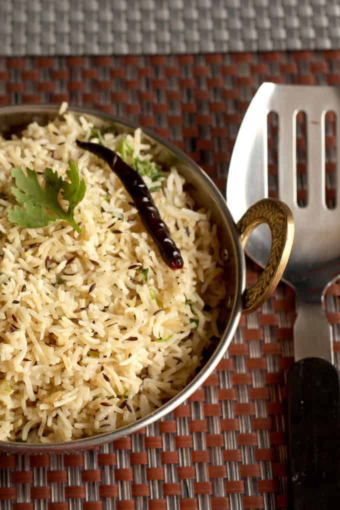 Fried jeera rice in a heavy vessel putted on a dark mat with some garnishing of coriander leaves and fried red chilli on it|