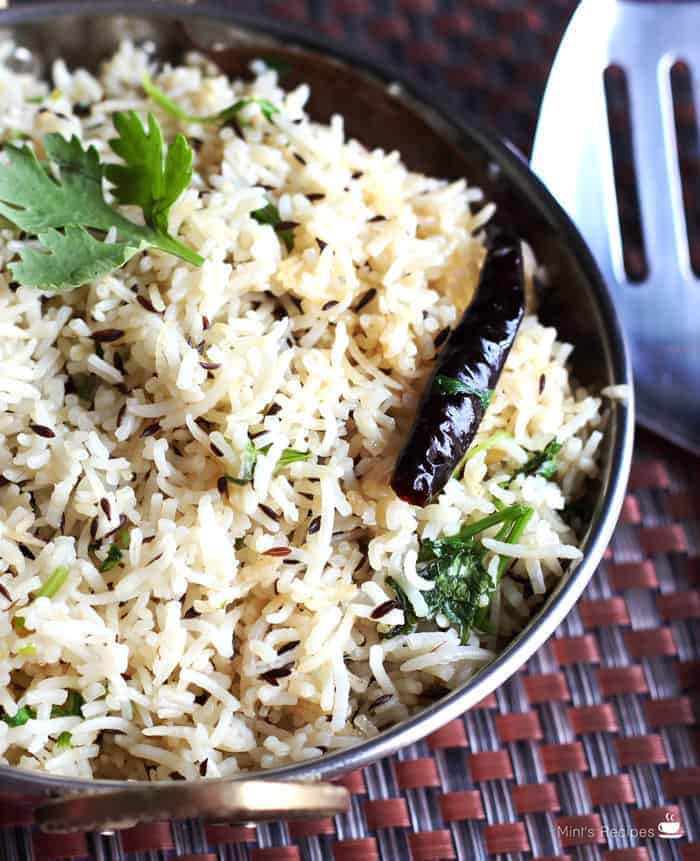 Fried jeera rice in a heavy vessel putted on a dark mat with some garnishing of coriander leaves and fried red chilli on it|