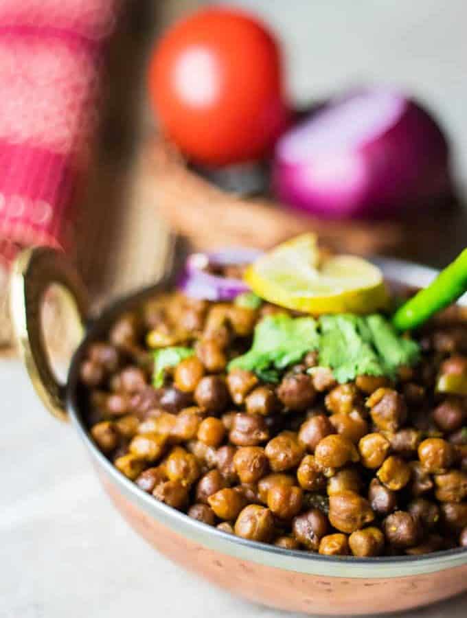 Dry kala chana on a deep vessel with coriander leaves and green chilli on top with a slice of lemon and some onion rings and light background with onion and tomato in a basket.