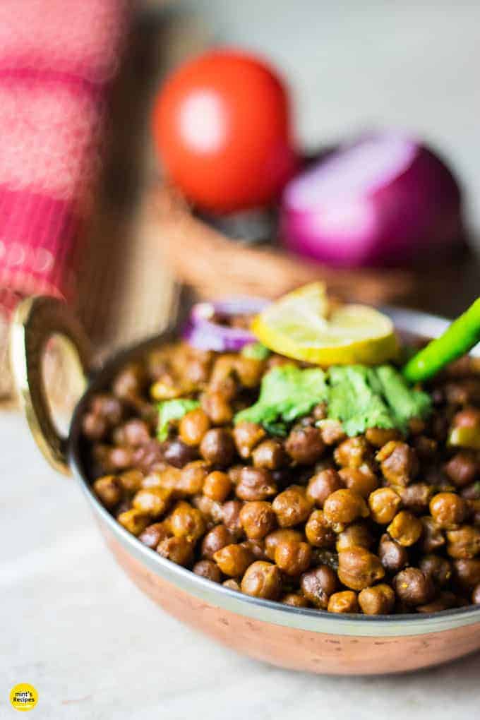 Dry kala chana on a deep vessel with coriander leaves and green chilli on top with a slice of lemon and some onion rings and light background with onion and tomato in a basket.