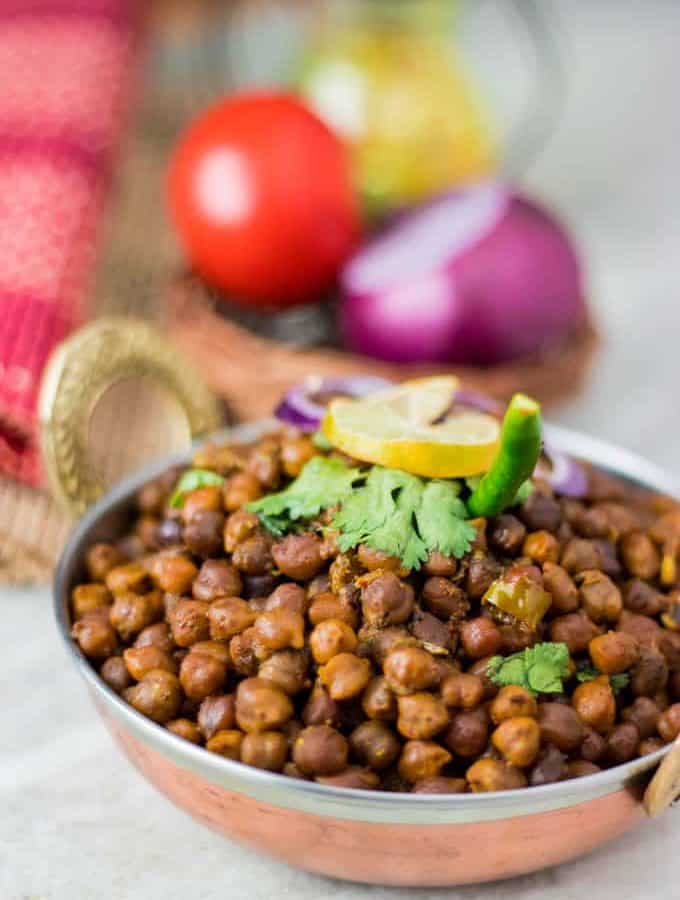 Dry kala chana on a deep vessel with coriander leaves and green chilli on top with a slice of lemon and some onion rings and light background with onion and tomato in a basket.