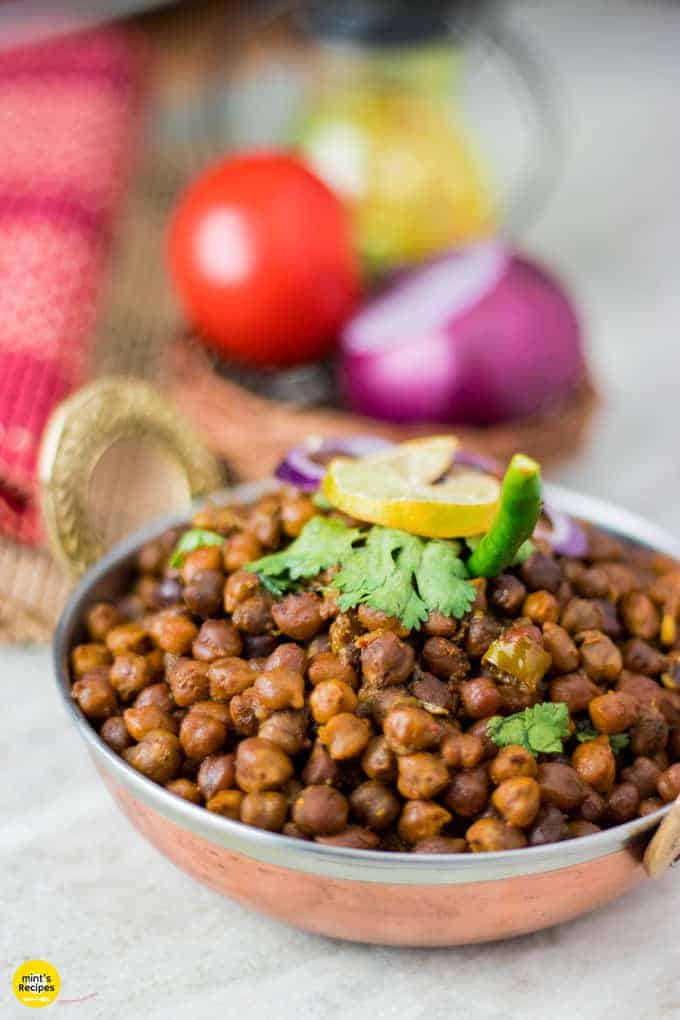 Dry kala chana on a deep vessel with coriander leaves and green chilli on top with a slice of lemon and some onion rings and light background with onion and tomato in a basket.