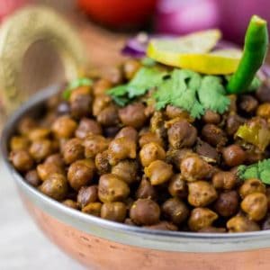 Dry kala chana on a deep vessel with coriander leaves and green chilli on top with a slice of lemon and some onion rings and light background with onion and tomato in a basket.