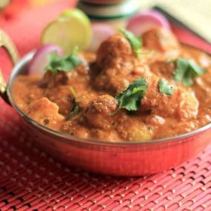 Kashmiri Dum Aloo on a deep vessel putted on a red mat garnished with some coriander leaves lemon wedges and some onion rings |