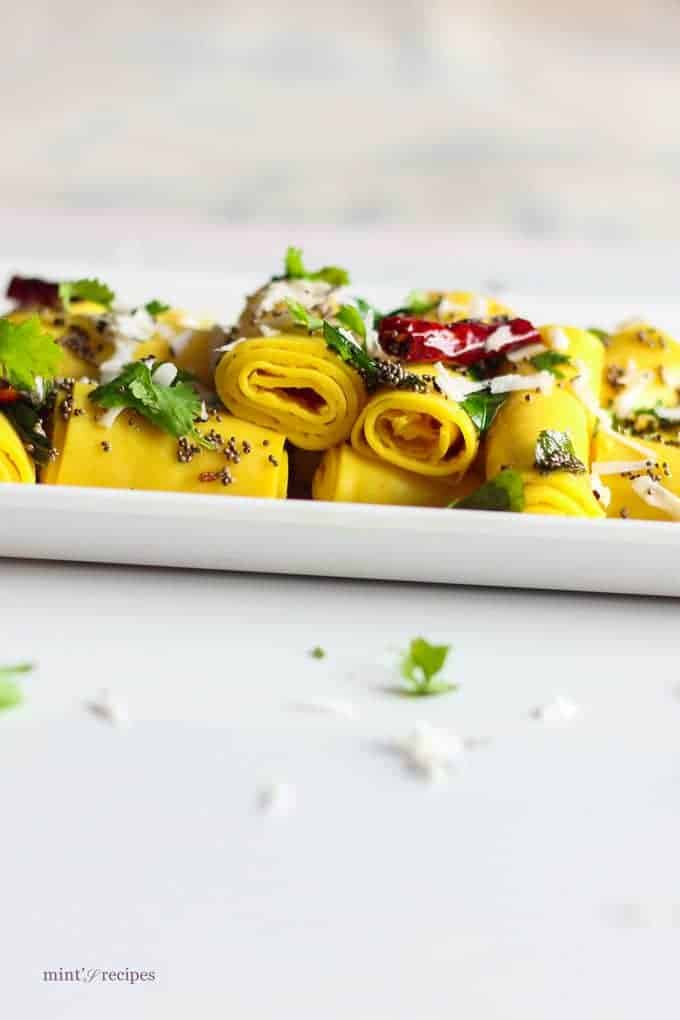 Khandvi on a white plate, garnished with some tempering of mustard seed and grated coconuts