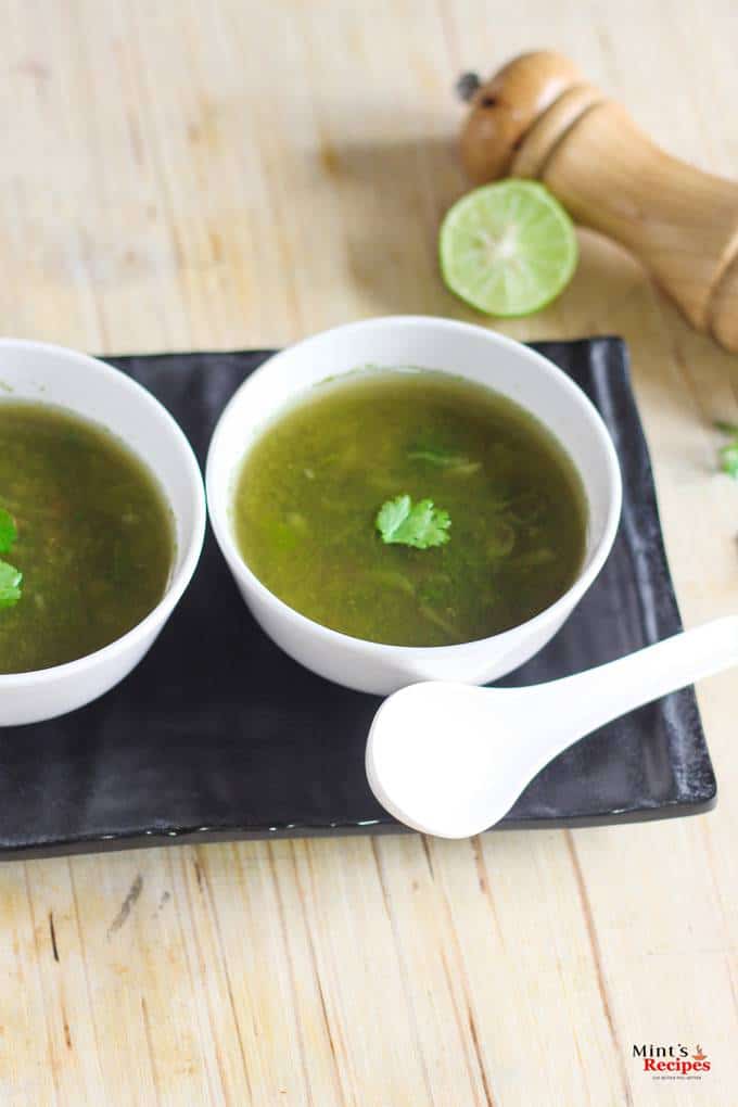 Lemon Coriander Soup on a white bowl kept on a black tray with a spoon and lemon slice and garnished with coriander leaves |
