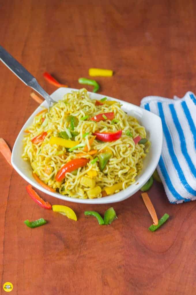 Masala maggi on a white bowl with lots of veggies spreaded around the bowl and a fork in it with a handkerchief on the side of the bowl |