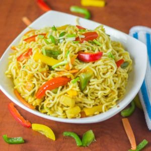 Masala maggi on a white bowl with lots of veggies spreaded around the bowl and a fork in it with a handkerchief on the side of the bowl |