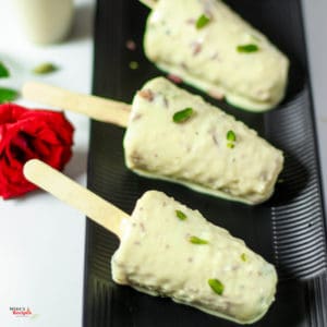 Malai kulfi stick on a black plate, kulfi coated with pista on a white light background with a red rose on the backside of the plate Roti noodles on a white bowl full with roti noodles and garnished with coriander leaves with a light background