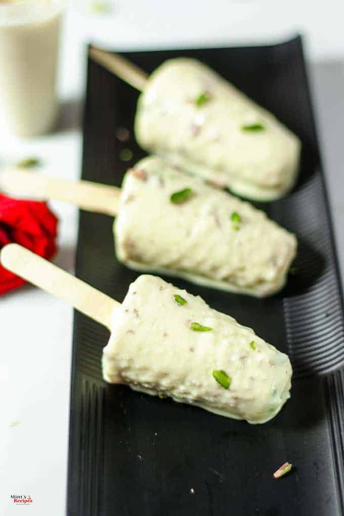 Malai kulfi stick on a black plate, kulfi coated with pista on a white light background with a red rose on the backside of the plate 