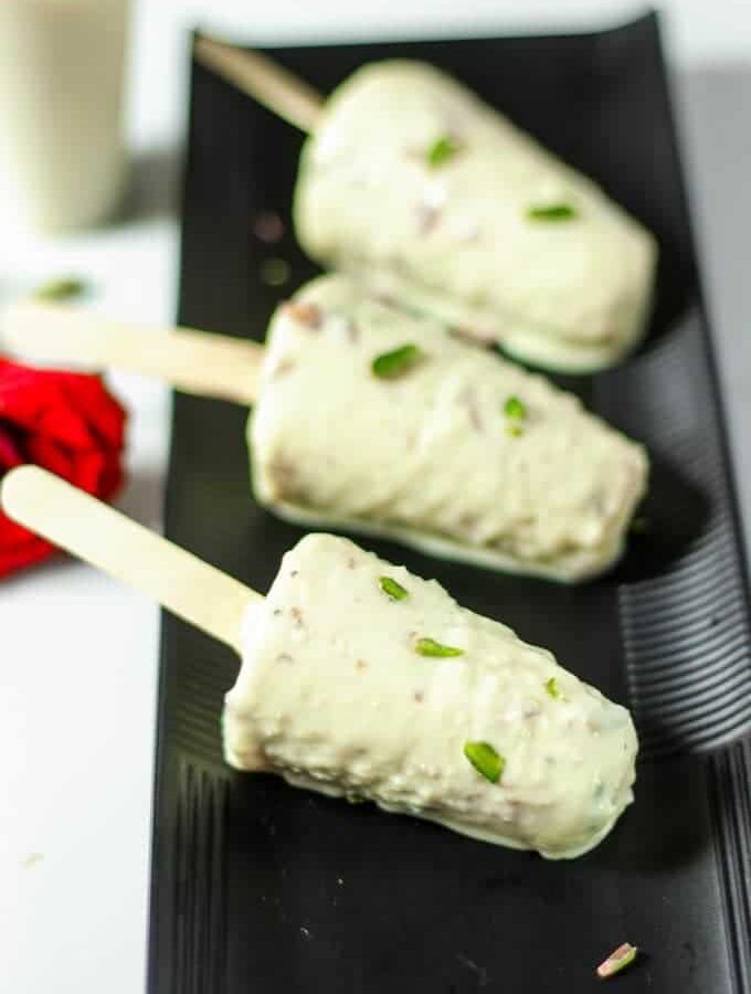 Malai kulfi stick on a black plate, kulfi coated with pista on a white light background with a red rose on the backside of the plate Roti noodles on a white bowl full with roti noodles and garnished with coriander leaves with a light background