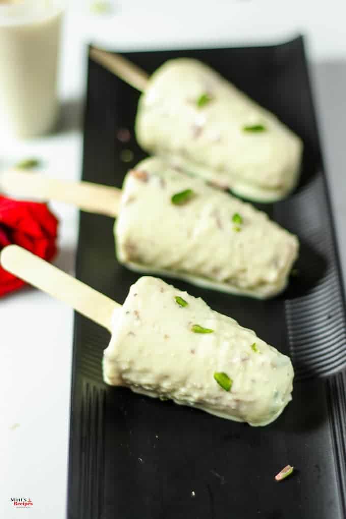 malai kulfi stick on a black plate. kulfi coated with pista on a white light background with a red rose on the backside of the plate |