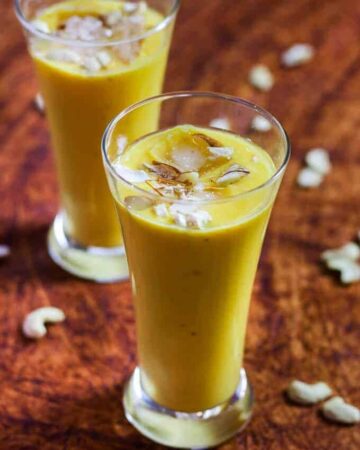 Mango Banana Smoothie on a glass with mango smoothie with some ice cubes, garnished with some cashew, almonds and saffron |