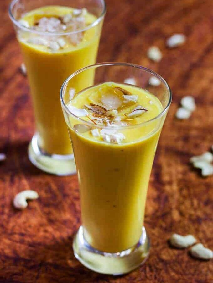 Mango Banana Smoothie on a glass with mango smoothie with some ice cubes, garnished with some cashew, almonds and saffron |