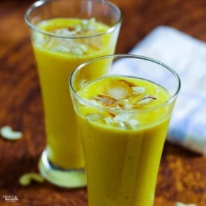 Mango Banana Smoothie on a glass with mango smoothie with some ice cubes, garnished with some cashew, almonds and saffron |