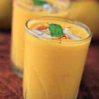 Mango Lassi on a glass with some ice cubes and some saffron with a mint leaf on it|