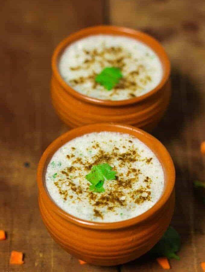 Masala Chaas in a clay pot with some cumin powder and coriander leaves and another masala chaas in the background kept on a wooden surface with some coriander leaves and chopped carrots to decorate the surface |