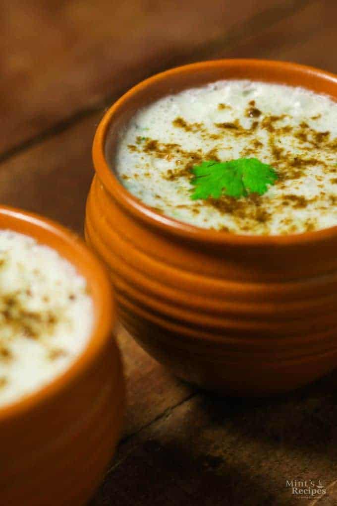 Masala Chaas in a clay pot with some cumin powder and coriander leaves and another masala chaas in the background kept on a wooden surface with some coriander leaves and chopped carrots to decorate the surface |