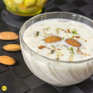 Meetha daliya kheer on a transparent small bowl full of daliya kheer and garnished with some chopped pista and almonds kept on a dark color surface with an decoration props on the background