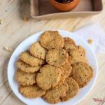 Methi Mathri on a white plate with some mathri stacked one another with some mathri on the sides of it and some background props