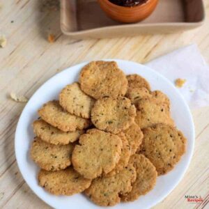 Methi Mathri on a white plate with some mathri stacked one another with some mathri on the sides of it and some background props